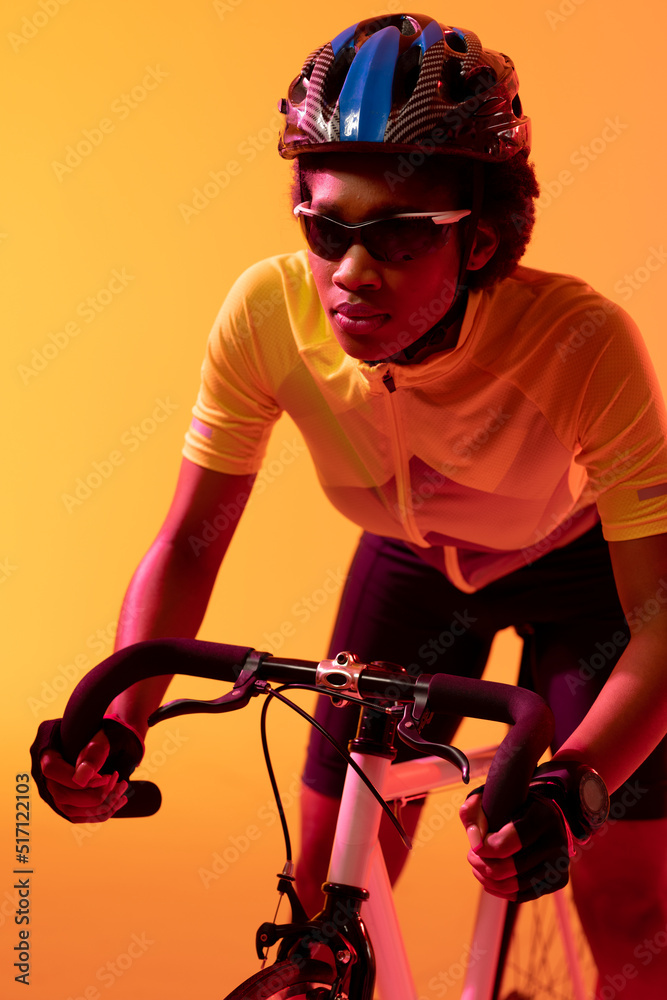 Vertical image of african american female cyclist riding bike in neon orange lighting