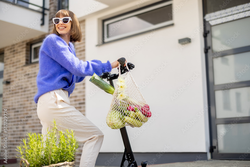 Happy stylish woman going home with fresh vegetables in mesh bag, driving on electrical scooter at r