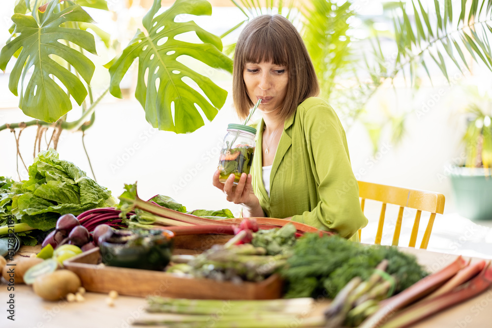 一位年轻女士坐在装满新鲜蔬菜、水果和gre的桌子旁，用瓶子喝水