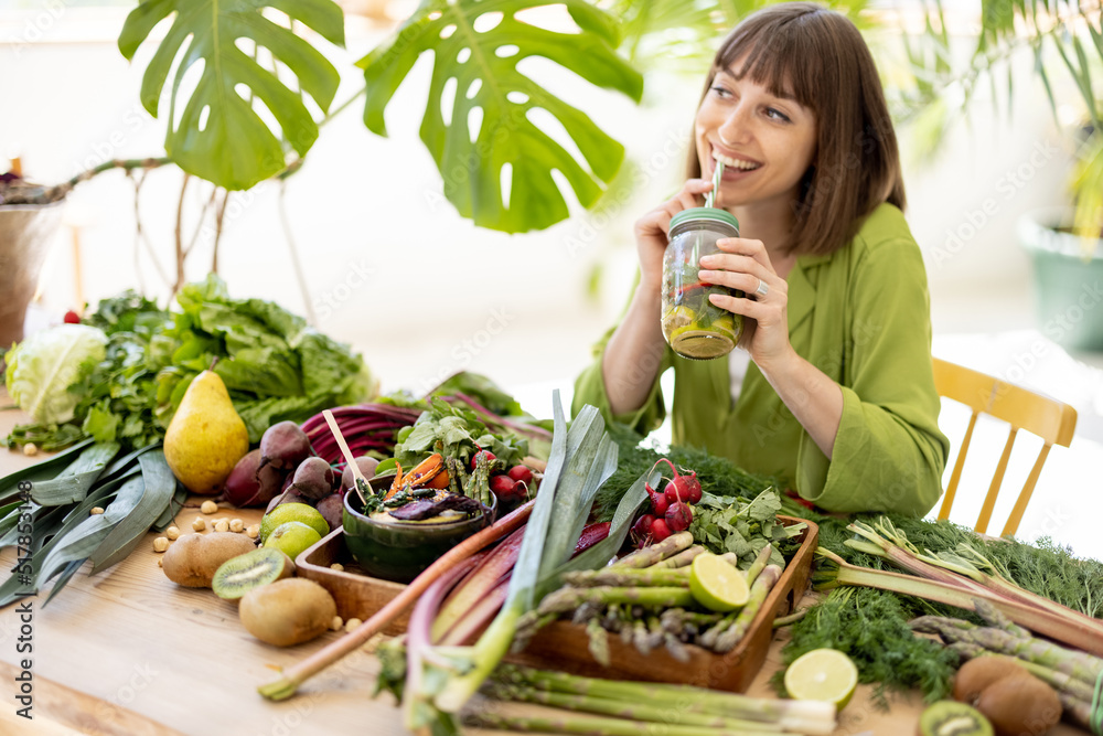 Young woman drinks from a bottle while sitting by the table full of fresh vegetables, fruits and gre