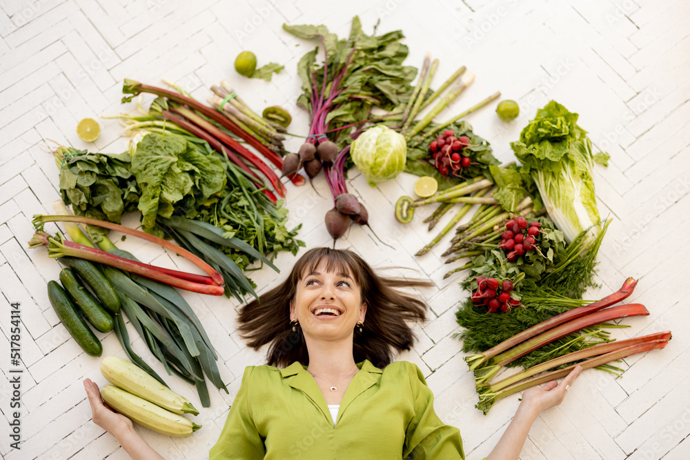 Portrait of a young cheerful woman with lots of fresh vegetables, fruits and greens above her head, 