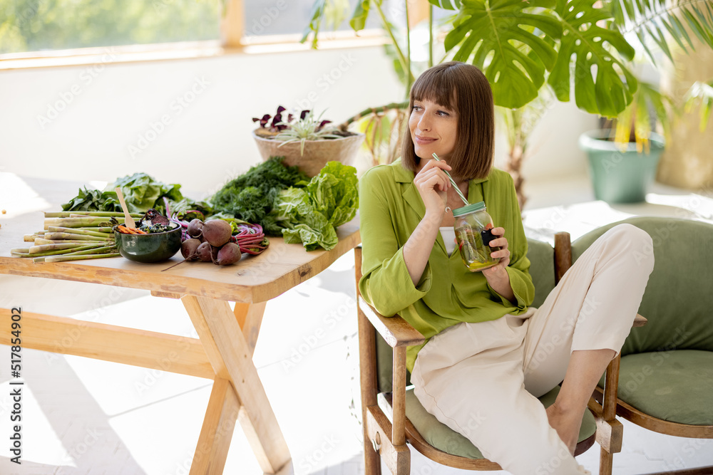 Young woman drinks lemonade while sitting on chair near table with lots of fresh food ingredients in
