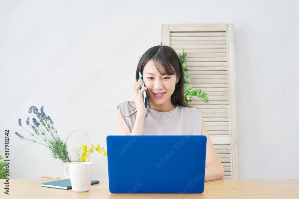 リビングでノートパソコンを見ながらスマホで電話する女性