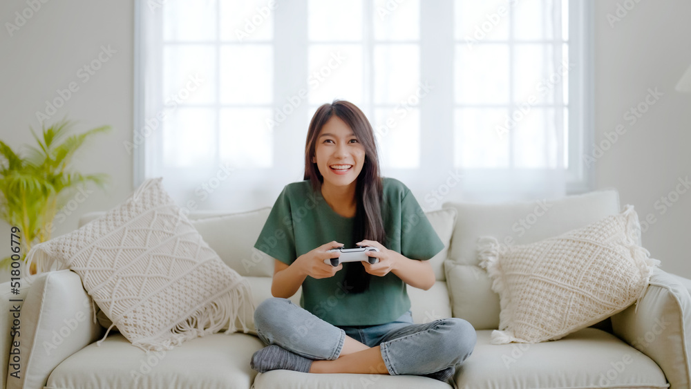 Beautiful young asian woman sitting in living room sofa holding joystick remote control playing onli