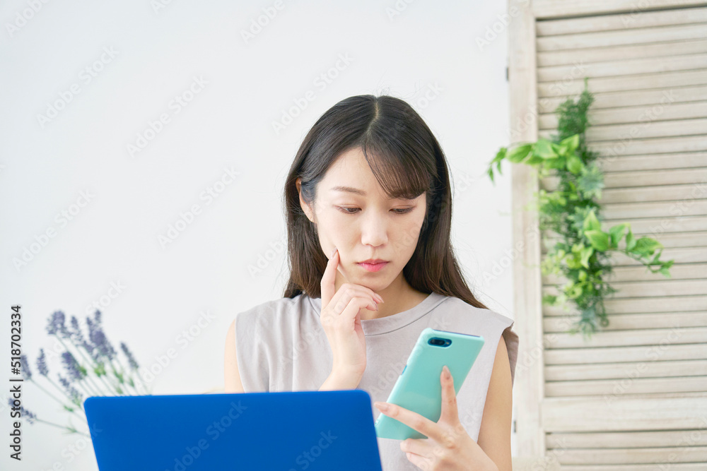 リビングでノートパソコンの前でスマホを見て考える女性