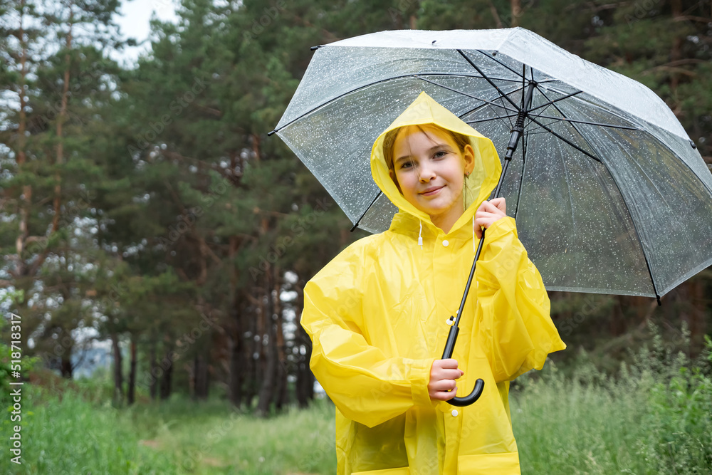 Smiling teenager girl stands against green forest in rainy weather. Young lady dressed in yellow rai