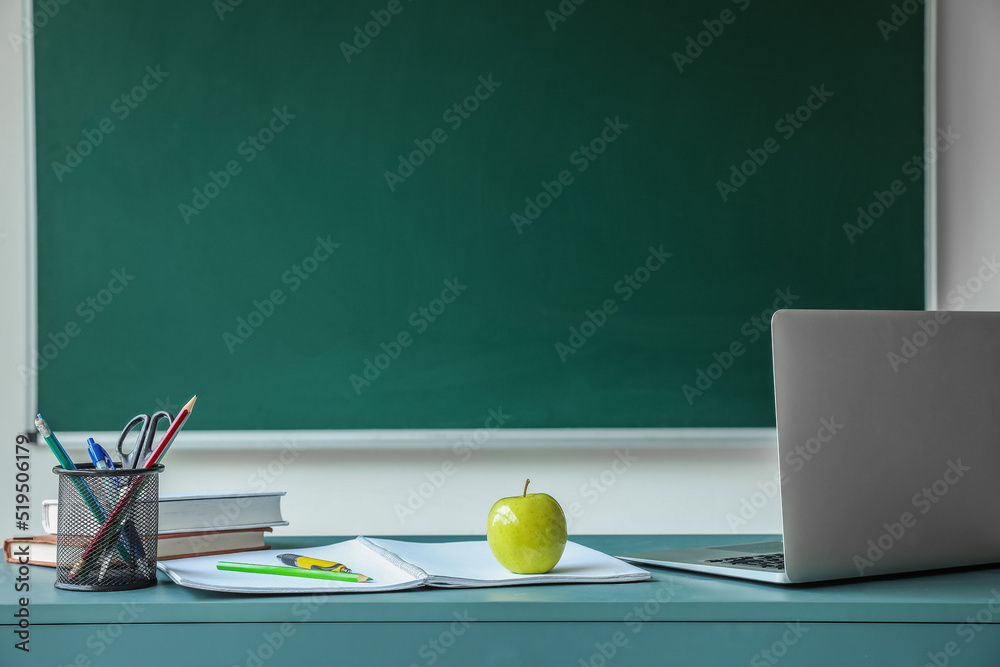 学校黑板附近的桌子上放着书、笔杯和笔记本电脑的苹果
