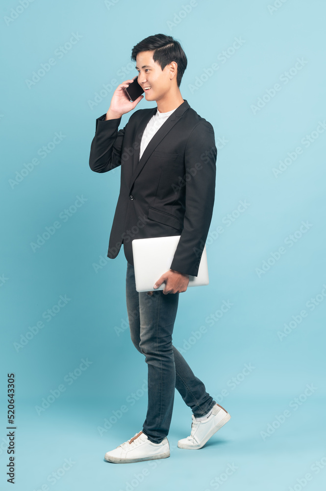 年轻的亚洲商人，穿着时髦休闲的风格，使用智能手机，拿着一台蓝色隔离的笔记本电脑