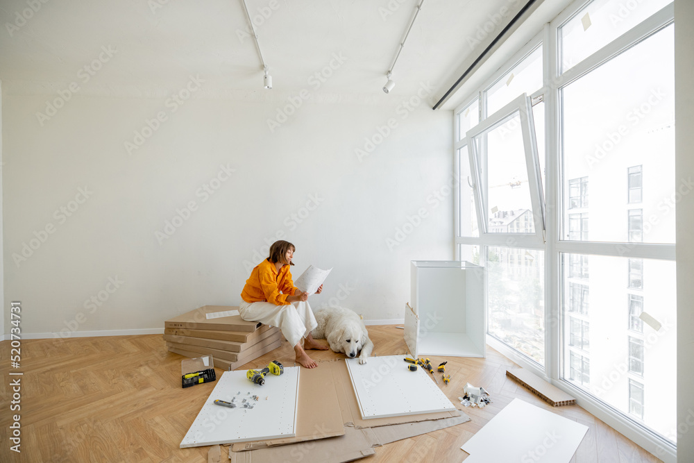 Young woman assembles furniture by herself, sitting with dog near window and reading manual at new a