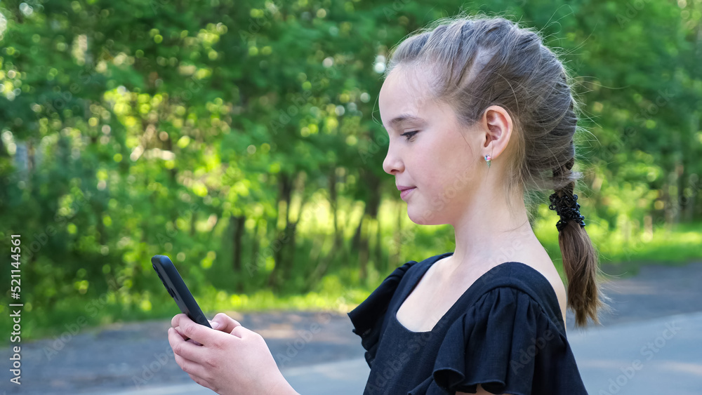 Schoolgirl with braid types on phone smiling. Stylish girl looks on phone screen standing on forest 