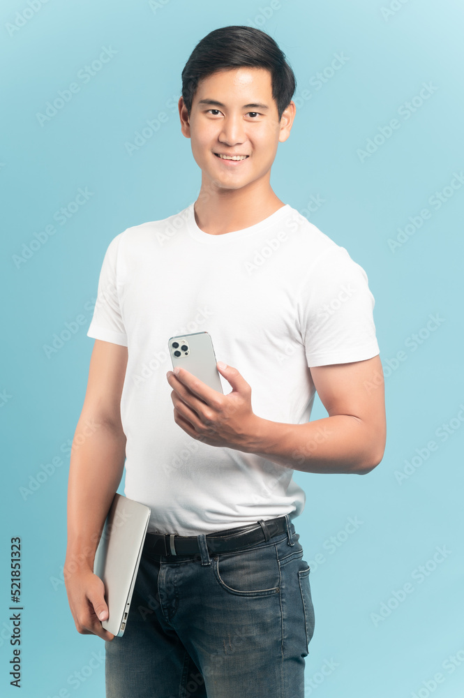 漂亮的亚洲人，穿着白色t恤和牛仔裤，手里拿着蓝色背景的智能手机和笔记本电脑