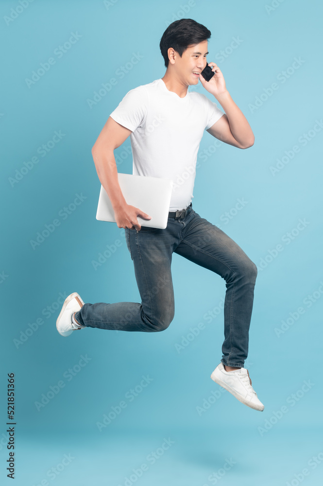 一个穿着白色t恤和牛仔裤的漂亮亚洲男人，手里拿着智能手机和笔记本电脑，在蓝色的bac上独自跳跃