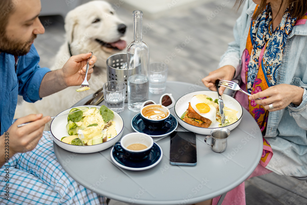时尚的男人和女人吃着美味的早餐，和可爱的狗坐在咖啡馆露台上。朋友们一起消费