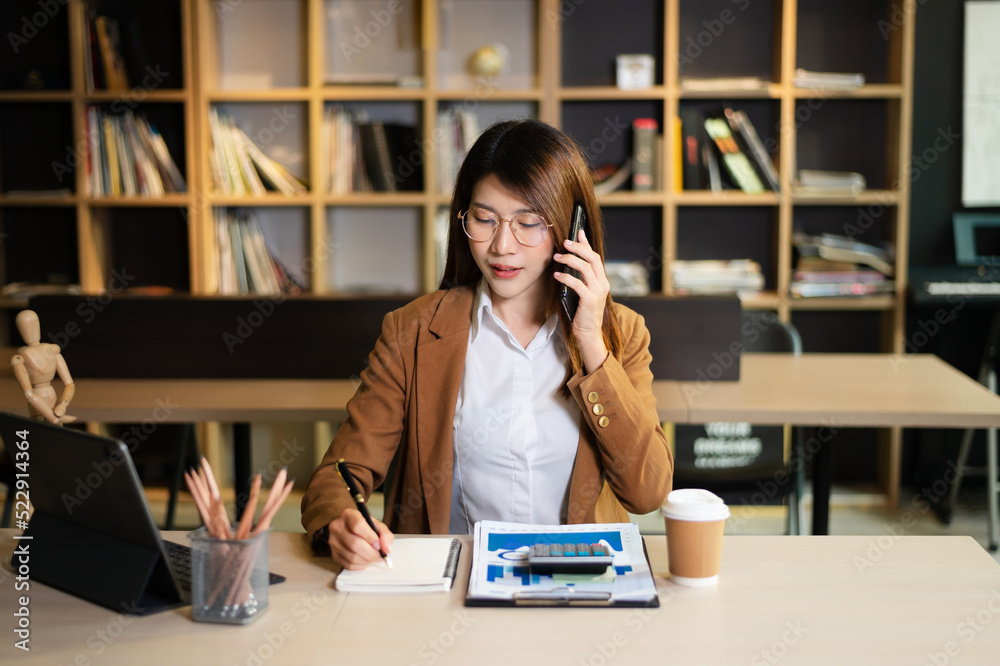 亚洲商务女性坐在办公室里微笑着打电话和使用笔记本电脑。