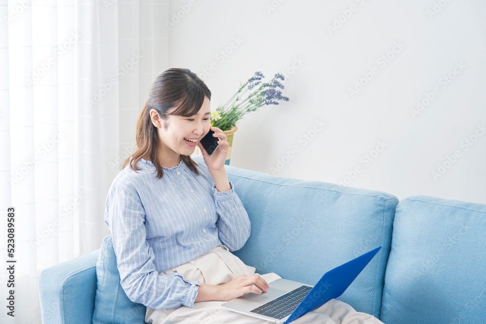 自宅でノートパソコンを見てスマホで電話する女性