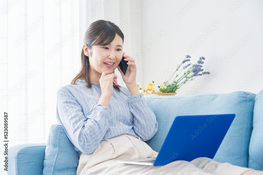 自宅でノートパソコンを見てスマホで電話する女性