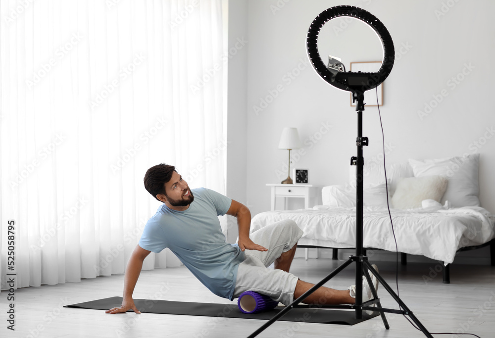 Young man with foam roller recording video in bedroom