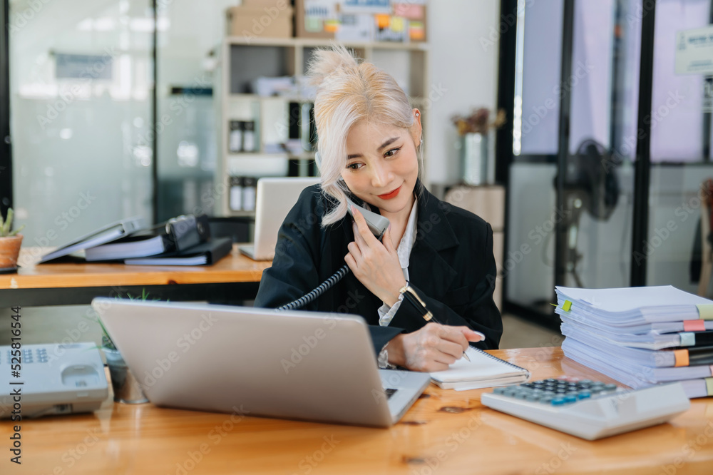 亚洲商务女性坐在办公室微笑着打电话和使用笔记本电脑