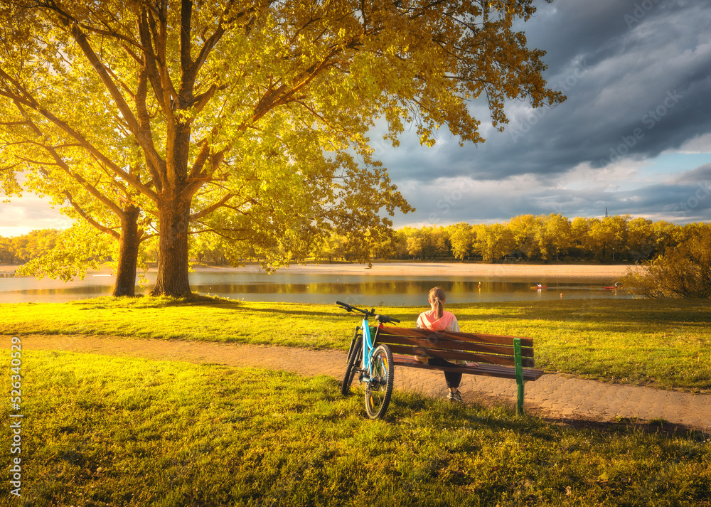 坐在长椅和山地自行车上的女人，黄色的树和湖