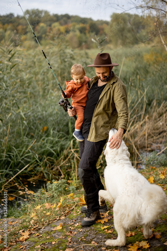 微笑的高加索男子带着小儿子和狗在河边或湖边钓鱼。休闲和