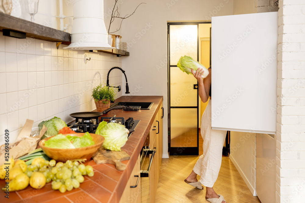 一位年轻女士用绿色新鲜食材烹饪健康素食，看着工具箱里的冰箱