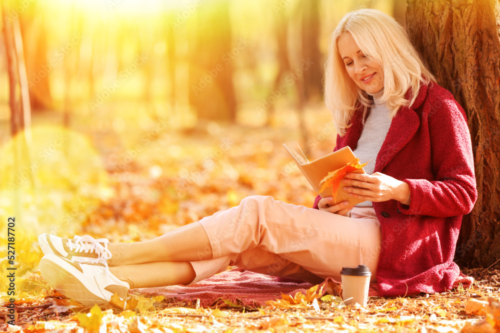 成熟的女人在秋天公园的树旁看书