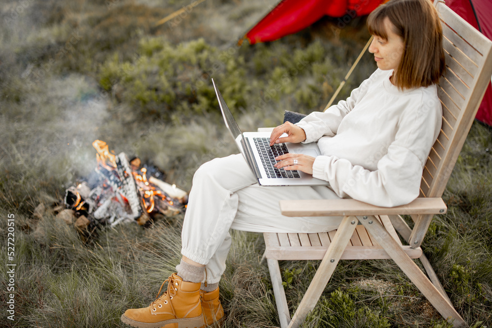 一名年轻女子在笔记本电脑上工作，放松地坐在篝火旁的椅子上，带着帐篷旅行