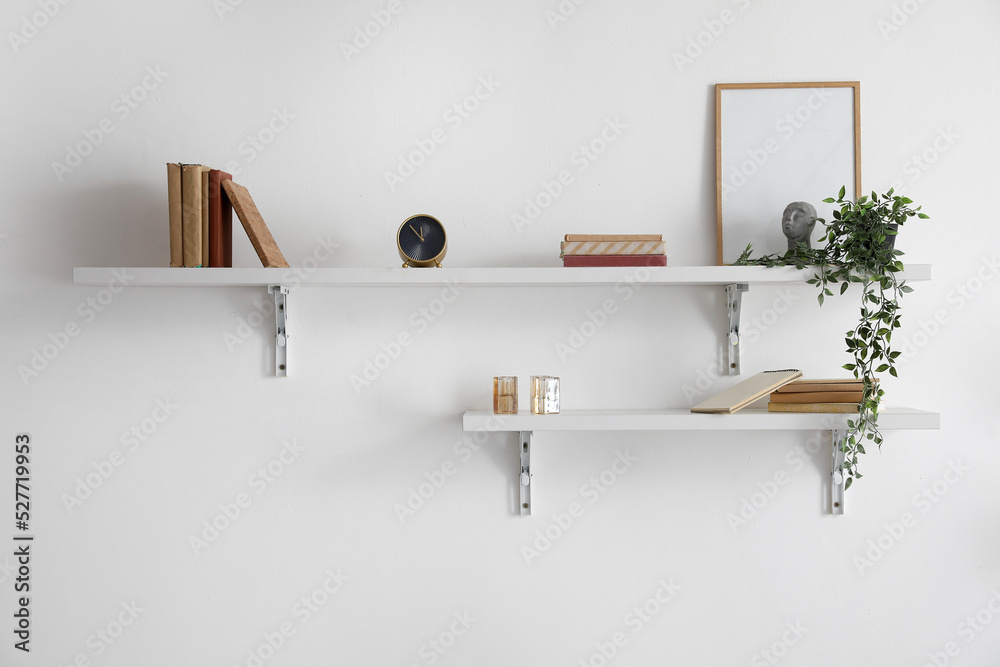 Shelves with books, frame, alarm clock and houseplant hanging on light wall