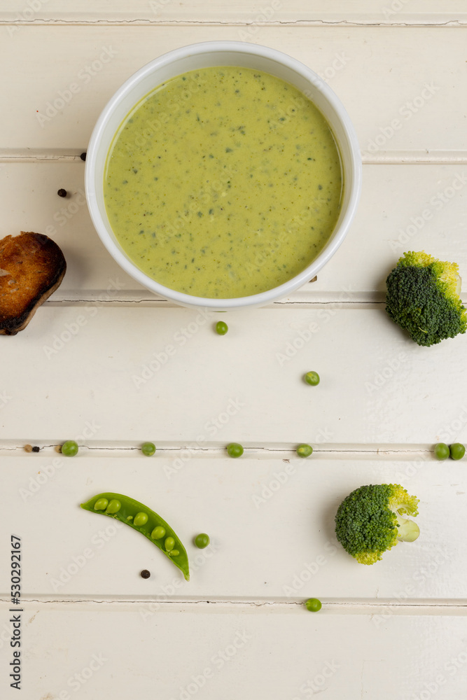 Vertical image of bowl of broccoli soup, with florets, peas and toast on white wood, copy space