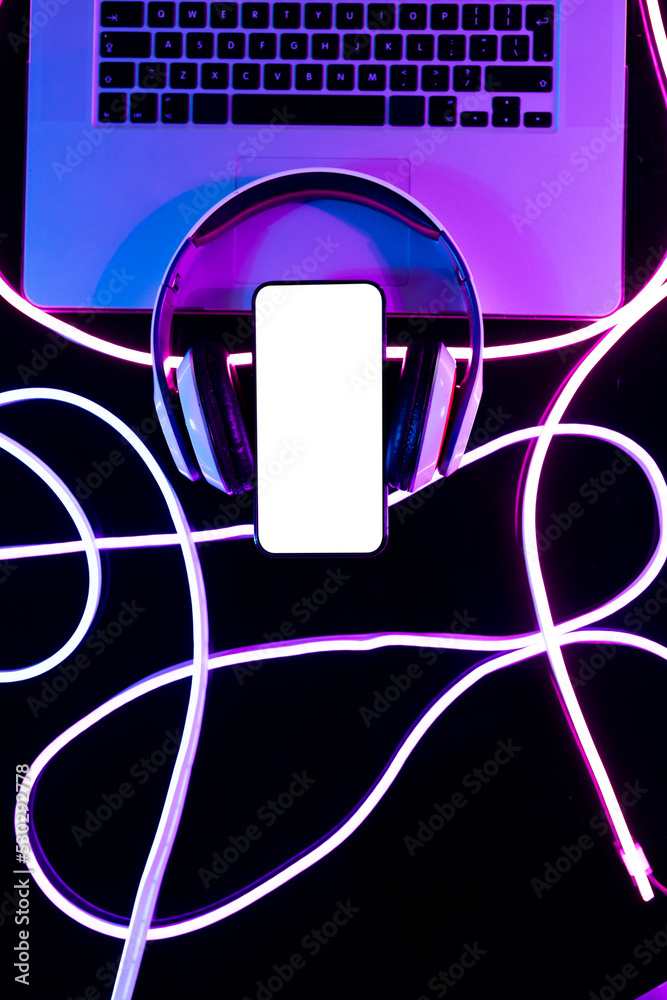 Image of headphones, laptop and smartphone with copy space over neon strands in background