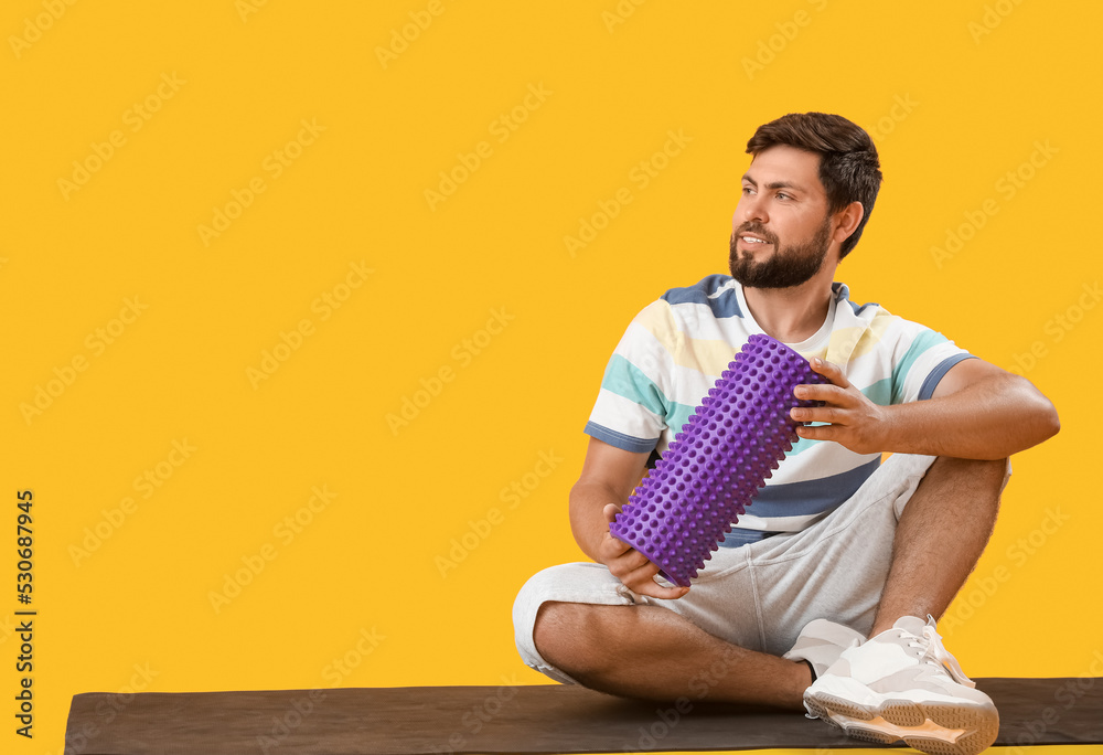 Young man with foam roller sitting on yellow background