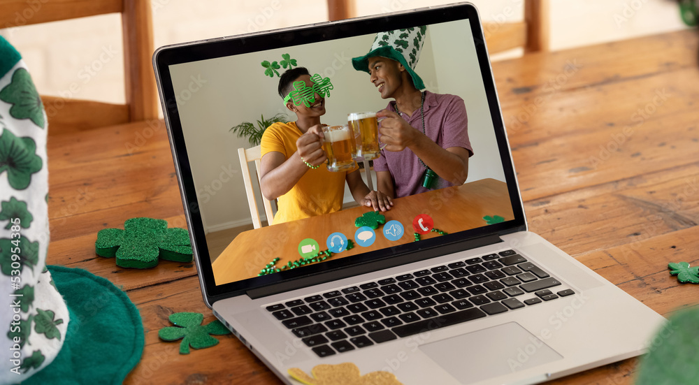 Webcam view of two african american men toasting beers on video call on laptop on wooden table