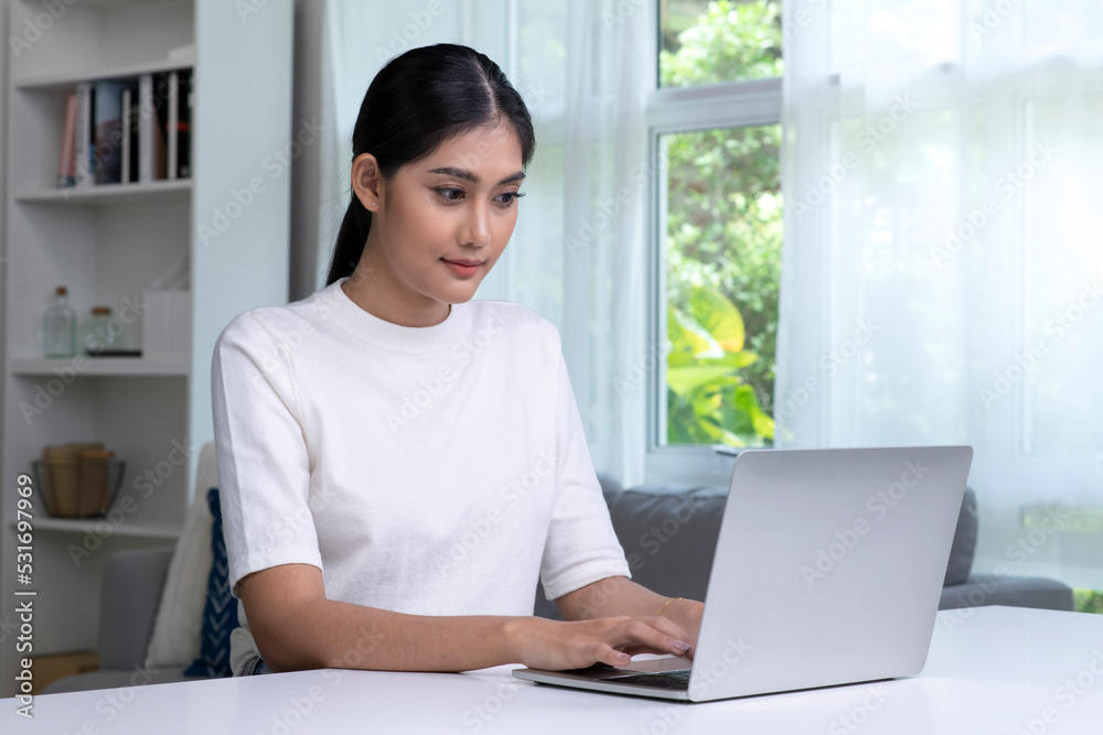 年轻的亚洲女性在家里使用笔记本电脑，看屏幕，聊天，阅读或写电子邮件，坐着