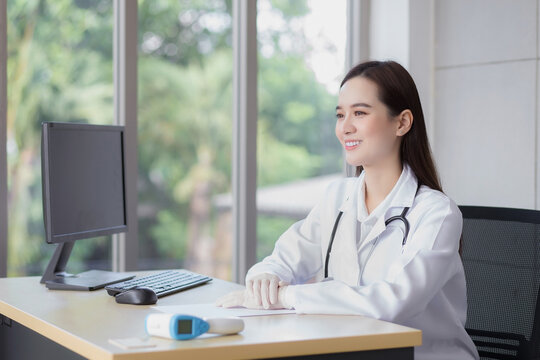 Asian professional young smiling woman doctor sitting look forward in office at hospital. On table has a paper and computer.