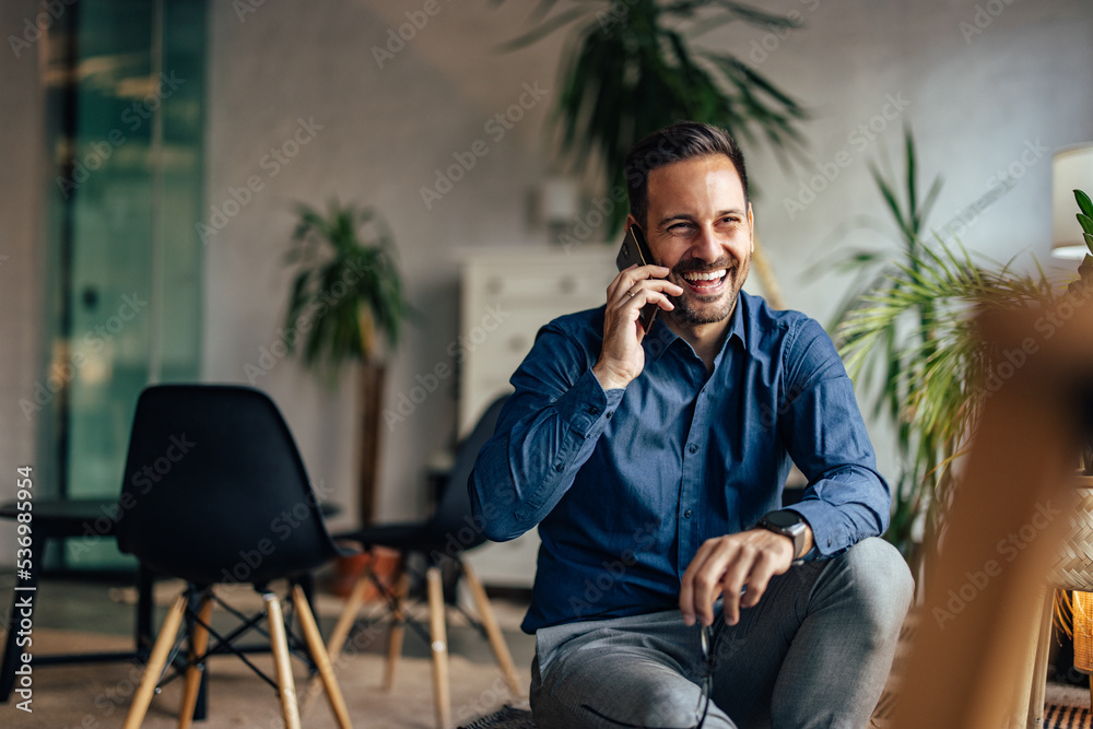 一个微笑的男人坐在室内，在办公室打电话。