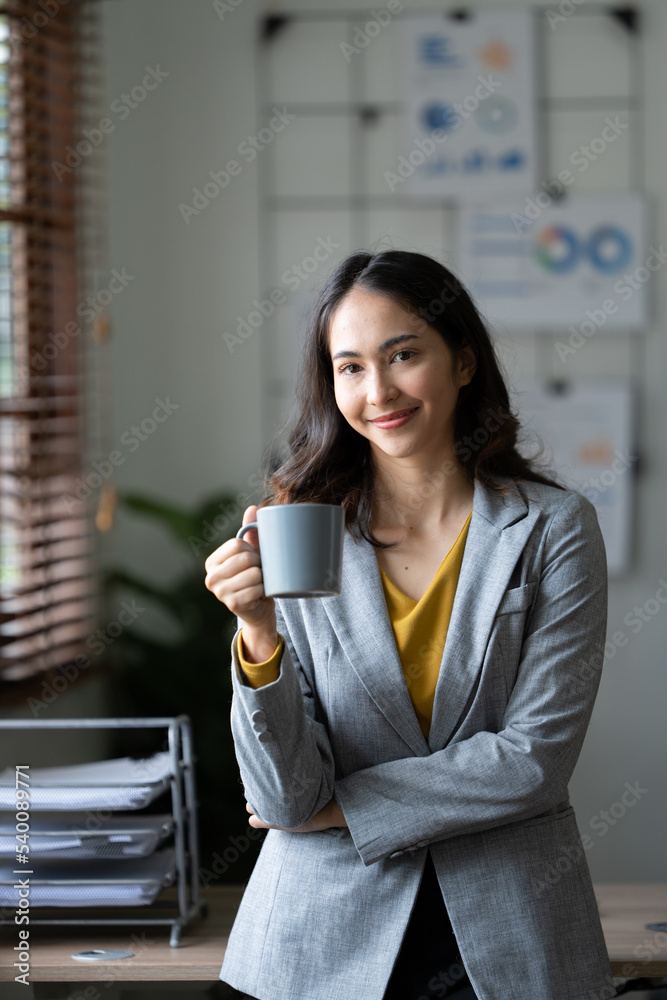 美丽的年轻亚洲女商人拿着咖啡杯和笔记本电脑在办公室工作，面带微笑。