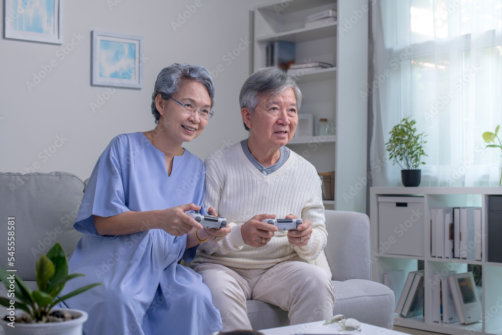 幸福的亚洲老年夫妇坐在客厅沙发上，手里拿着控制器，大笑
