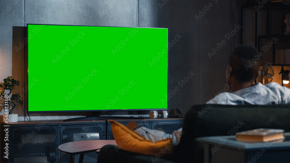 黑人坐在家里的沙发上观看带Chromacey的绿屏模拟电视显示屏。R