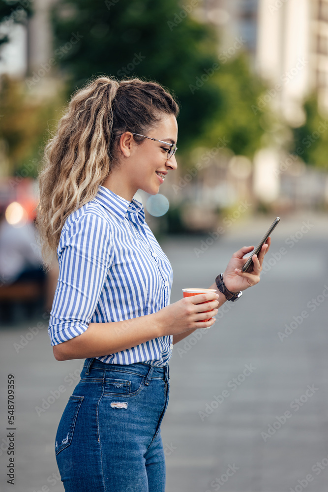 一个面带微笑的女孩站在户外，手里拿着手机，手里喝着咖啡的侧视图。