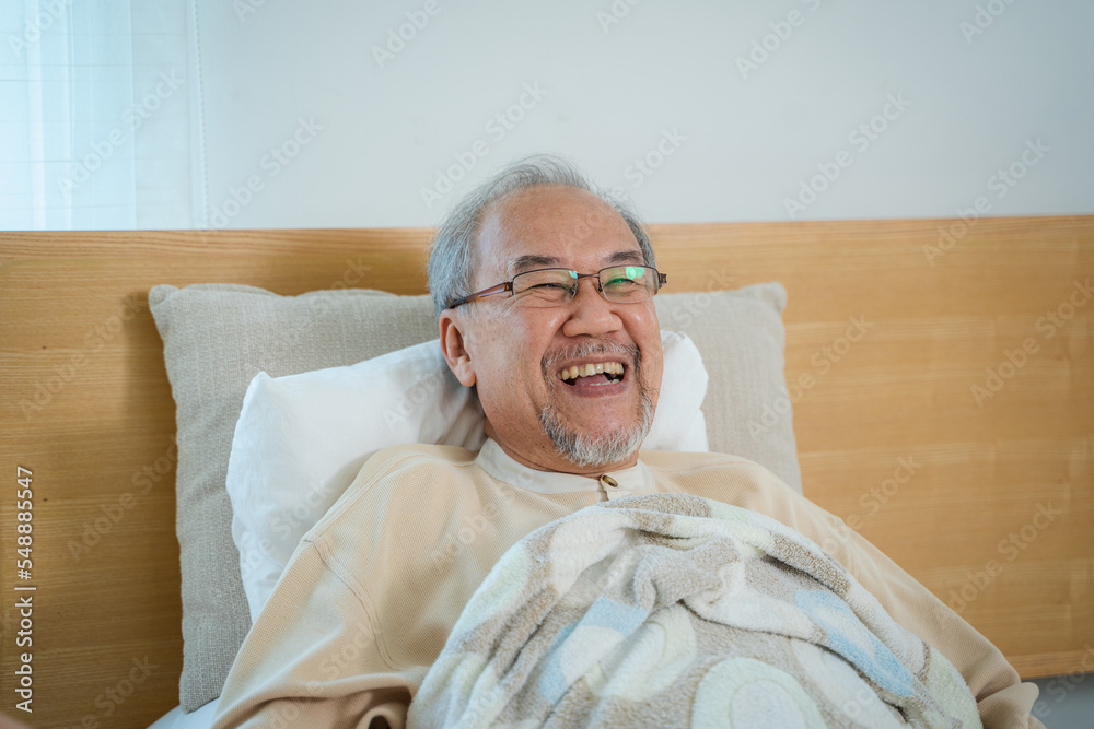 躺在家里卧室的床上，一位亚洲老人的特写脸正开心地笑着。