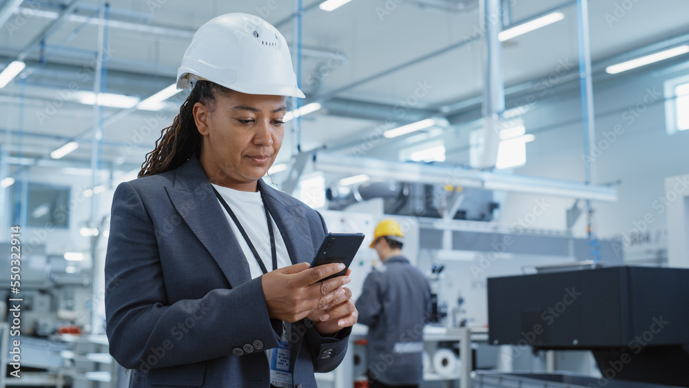 Portrait of a Black Female Engineer in Hard Hat Standing and Using a Smartphone at Electronics Manuf