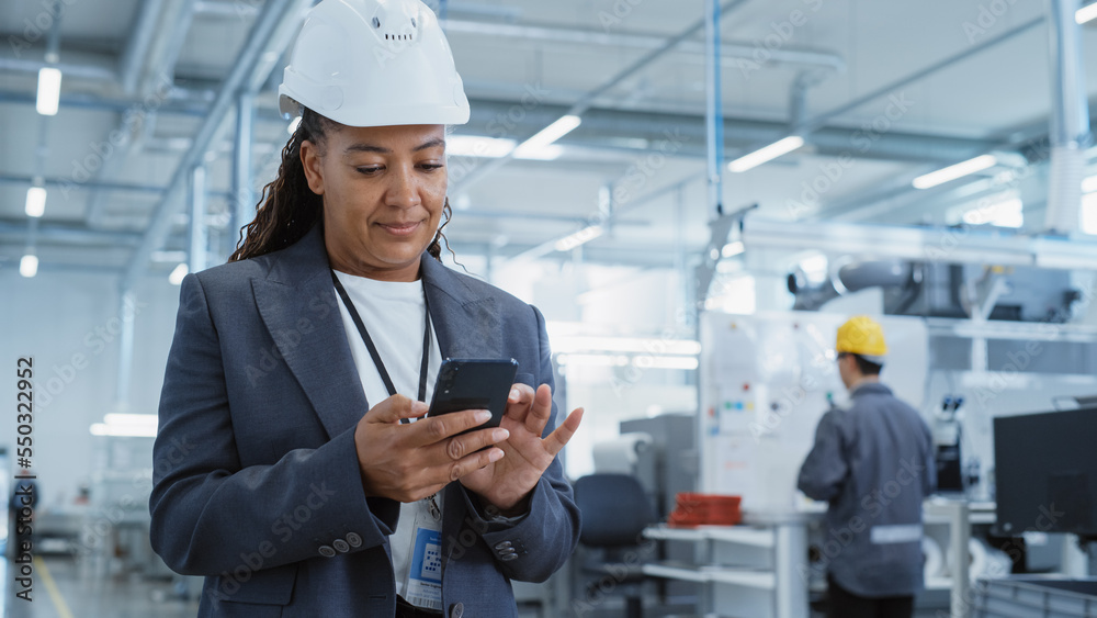 戴着安全帽的黑人女工程师在电子制造厂站着使用智能手机的肖像