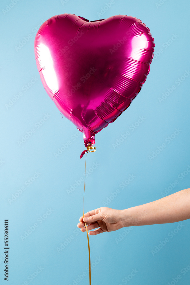 手持闪亮的粉红色心形气球的垂直图像，漂浮在蓝色背景上，带有复制空间