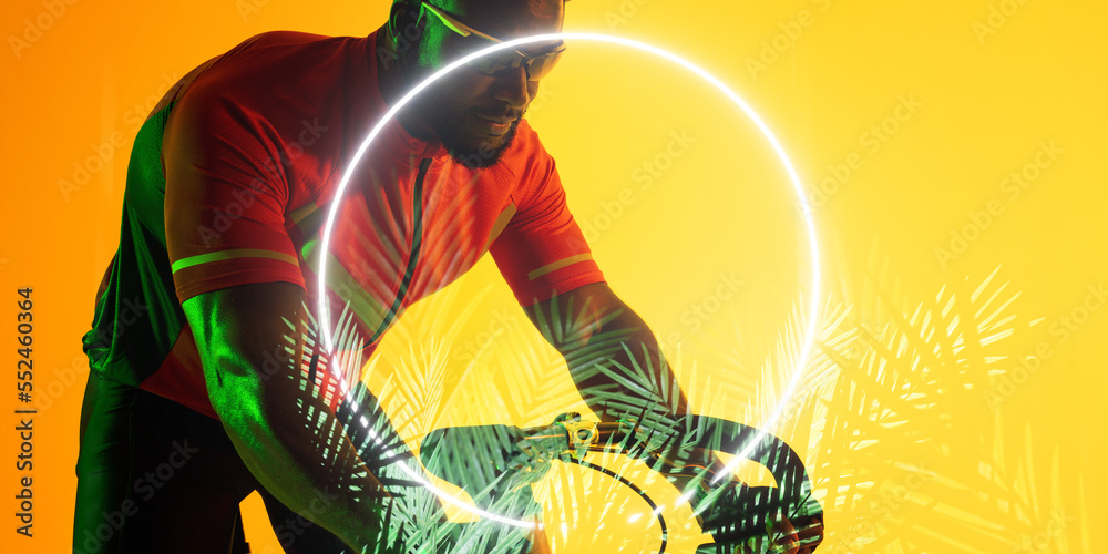 African american male athlete riding bike by illuminated circle and plants on yellow background