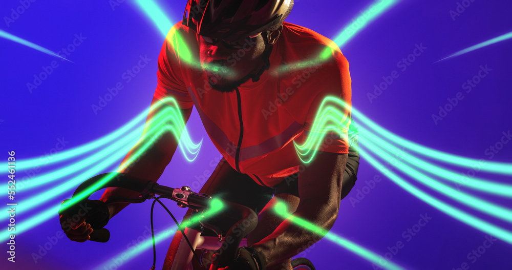 African american male athlete wearing helmet riding bike by illuminated lines on blue background