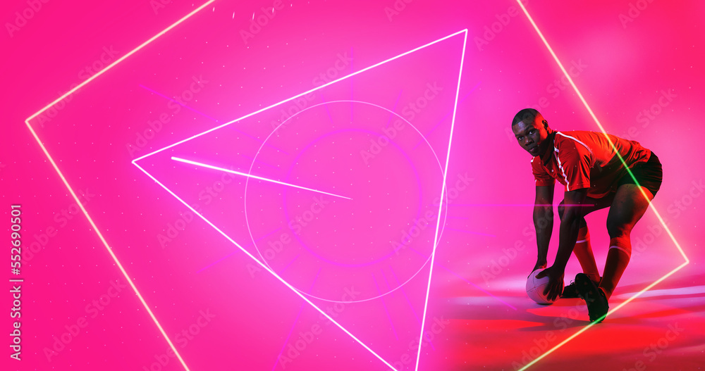 African american rugby player bending and taking ball over geometric shapes on pink background