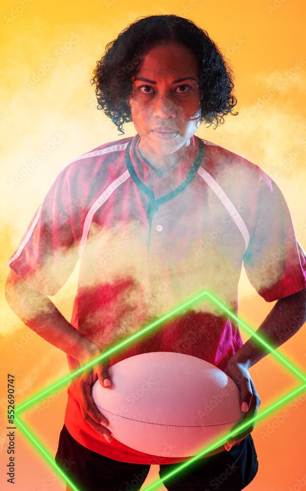 自信的混血女橄榄球运动员拿着球站在烟雾中，被照亮的长方形