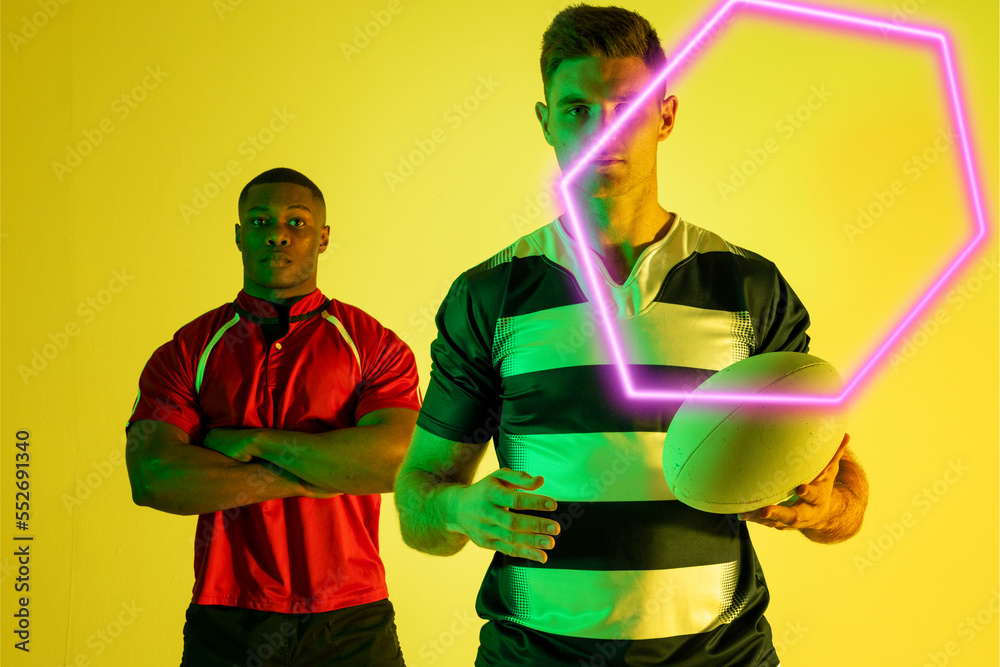 Diverse confident male rugby opponents standing with arms crossed and ball by illuminated hexagon