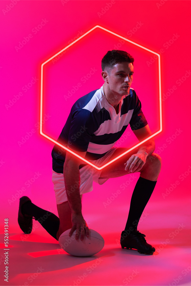 Caucasian male rugby player with ball kneeling by illuminated hexagon over pink background
