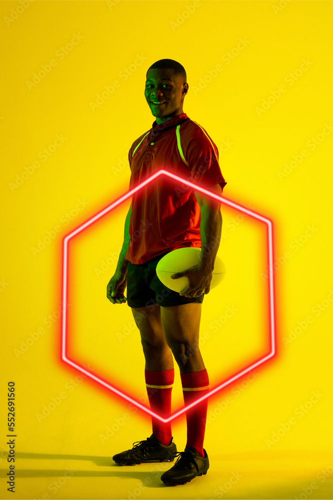 Smiling african american male player with rugby ball by illuminated hexagon on yellow background